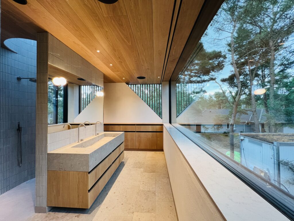 Exklusivt badrum i Villa P2 med väggar och inredning i trä och sten, stort fönster och takfönster i duschen som släpper in naturligt ljus. Harmonisk design med noggrant utvalda material och tidlös elegans.